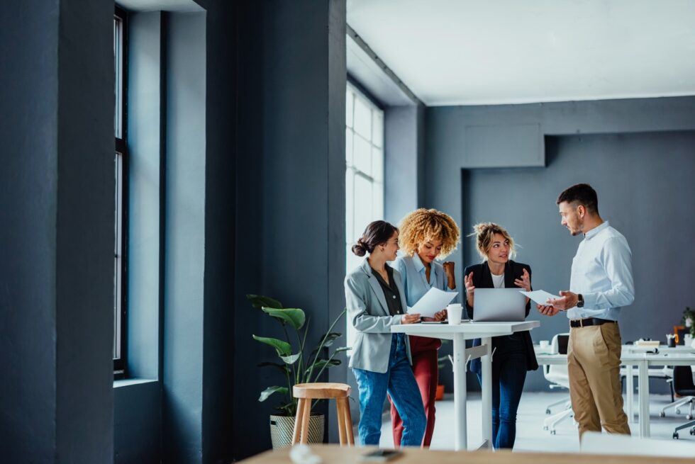 Multi ethnic team of four colleagues using laptop computer and business papers while discussing ideas during a casual meeting in the office.